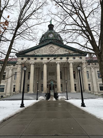 A stately, classical-style building stands prominently under an overcast, grey sky on a winter day in Binghamton, United States. The building, constructed from light-colored stone or brick, features a grand central facade with a portico supported by multiple tall, fluted columns, likely Ionic or Doric. Above these columns, a decorative pediment crowns the main entrance. Perched atop the building's central structure is a distinctive domed clock tower, appearing to be made of oxidized green copper, with multiple clock faces clearly visible, though the exact time is indiscernible. A dark statue on a pedestal is situated in front of the main entrance, nestled between two of the columns.
Bare, skeletal tree branches, characteristic of winter, frame the upper left and central portions of the image, with a scattering of dried brown leaves still clinging to some twigs. The foreground shows a concrete sidewalk running along the right side of the frame. Adjacent to the sidewalk, between it and the building, is a strip of ground covered in patches of snow and ice, emphasizing the cold weather. Several black, traditional-style street lamp posts are spaced along this snowy border, lining the approach to the building. The overall scene depicts a quiet, cold, and overcast winter day.