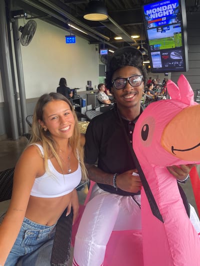 Two individuals are smiling directly at the camera in what appears to be a Topgolf facility located in Oxon Hill, United States. The female on the left has light brown hair and is wearing a white ribbed crop top and light blue jeans, adorned with two necklaces and small hoop earrings. The male on the right, with dark curly hair, black-framed glasses, and a nose piercing, is wearing a black polo shirt and light-colored pants, while holding a large, inflatable pink flamingo.
The background reveals a multi-level golf driving range setting with various patrons and staff. Digital display screens show bay numbers "208" and "207." A large overhead monitor prominently displays "TOPGOLF" branding, "MONDAY NIGHT FOOTBALL" details (Cincinnati vs. TBD, dated 09.29.25), golf game statistics, and "APEX CALLAWAY" logos. Overhead fans and lighting fixtures are visible. The floor to the right features a red stripe.