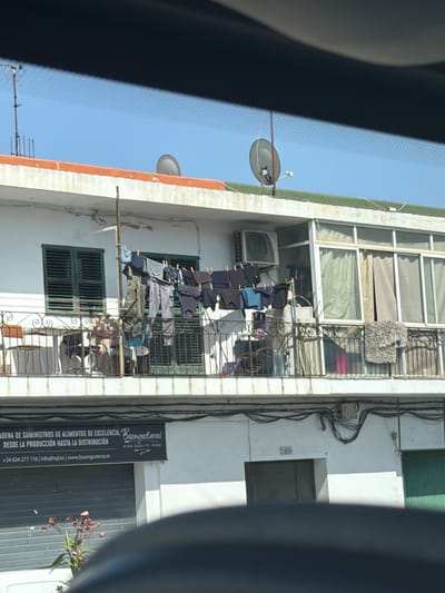This image shows a building with a balcony in Can Negre, Spain. The balcony has laundry hanging on a clothesline. There are also some chairs and a table on the balcony. In the foreground, there is a glimpse of a car's interior, suggesting the photo was taken from a vehicle. Below the balcony, a storefront with a sign reading "CADENA DE SUMINISTROS DE ALIMENTOS DE EXCELENCIA. Buongusterai HIGH QUALITY FOOD" is visible. The sign also includes a phone number and website. Further down, there are two doors, one labeled with the number 2400. A small plant with pink flowers is also visible in the lower left corner. The sky is clear and blue.