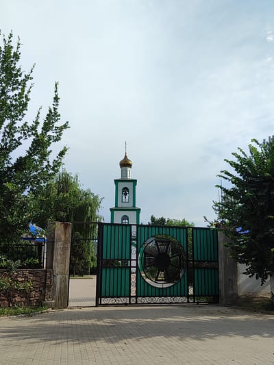 A daytime, eye-level, medium shot captures a view through an open, ornate green metal gate. The gate features decorative scrollwork and a circular motif in its center. Beyond the gate, a paved pathway leads to a church bell tower with a golden dome. The tower is white with green accents and has a bell visible through an arched opening. Lush green trees flank the scene, with branches reaching towards a bright, overcast sky. The ground in the foreground is paved with light-colored rectangular blocks, casting shadows. The image depicts a peaceful, serene scene, likely a religious site in Boraldai, Kazakhstan, during the day under cloudy skies. No people are visible, and there is no visible text in the image.