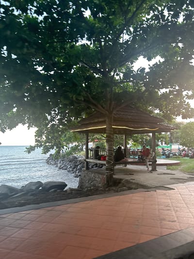 The image depicts a serene waterfront scene at Pantai Seruni, a popular tourist spot in Bantaeng, Indonesia. The foreground is dominated by a paved area with reddish-orange tiles, suggesting a walkway or seating area. To the left, the calm blue sea stretches towards the horizon, meeting a rocky shoreline.
A prominent feature of the scene is a rustic wooden gazebo situated along the coast, providing shade and a resting place for visitors. The gazebo is decorated with string lights, indicating it may be illuminated at night. Several individuals are visible inside and around the gazebo, with some seated and others standing. They appear to be enjoying the view or the company.
Behind the gazebo, a row of motorcycles is parked, suggesting that visitors typically arrive by private vehicle. Lush green trees and vegetation surround the area, adding to the tranquil atmosphere. The overall impression is one of a relaxed, coastal recreational area.