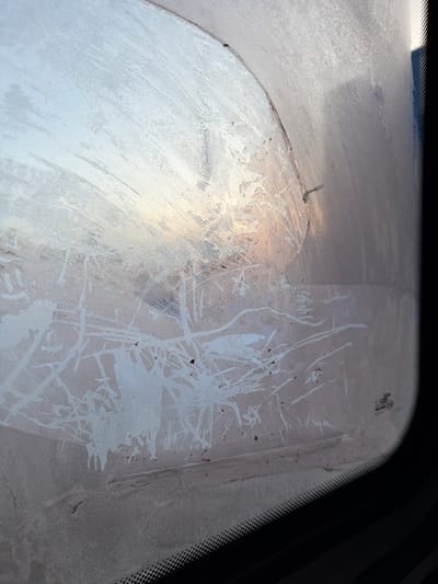 The image presents an interior perspective through a heavily frosted vehicle windshield, observed in the city of Золотая Степь, Russia. The glass is substantially obscured by dense, crystalline frost patterns. A partial, arced area in the upper-center, consistent with a windshield wiper's sweep, exhibits less frost, revealing a hazy exterior background with pale blue and faint orange-pink light, indicative of dawn or dusk. Below this cleared section, the frost features intricate, abstract branching and irregular linear formations, interspersed with scattered dark particulate debris. A small, dark, angular anomaly is visible near the top edge of the frosted area. The lower portion of the image displays the vehicle's black dotted frit band along the windshield's edge, and a faint manufacturer's emblem is discernible in the lower right corner of the glass. The overall scene suggests cold ambient temperatures.