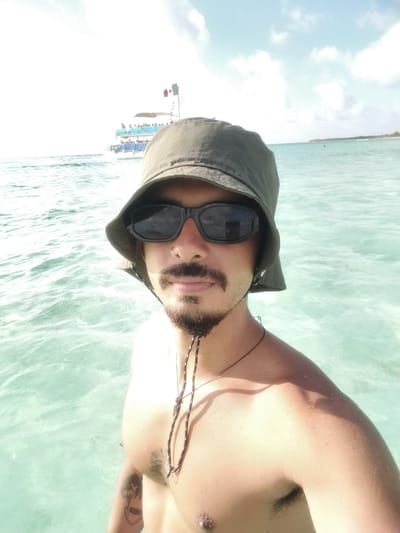 A person is standing in turquoise ocean water, likely off the coast of El Cedral, Mexico. The person is wearing a green bucket hat and dark sunglasses, with a mustache and goatee. They have a nose ring, necklace, and tattoos on their chest and left arm. The individual is shirtless and appears to be taking a selfie. In the background, a tour boat with people visible on the top deck is sailing on the water. The Mexican flag is visible on the boat. The sky is partly cloudy. In the distance, there is a hint of land.