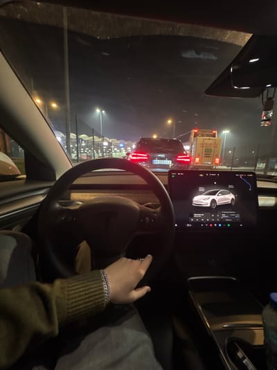 A nighttime view from inside a car, likely a Tesla, shows the driver's hands on the steering wheel. The car is in traffic, with other vehicles visible through the windshield. A large screen displays a graphic of a car and other data. The surroundings are dark, illuminated by streetlights and vehicle lights. The driver wears a long-sleeved shirt and a bracelet.