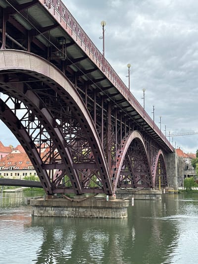 The image shows a close-up view of the Francis Bridge (Frančiškovem mostu) in Maribor, Slovenia, crossing the Drava River. The bridge is made of dark red metal and features a series of arched supports. The water of the Drava River reflects the bridge's structure, appearing greenish in color. The sky above is overcast with gray clouds. In the background, buildings with red-tiled roofs and lighter colored facades are visible on the riverbank, along with some greenery and a crane in the distance. The scene is static, with no people or immediate actions depicted.