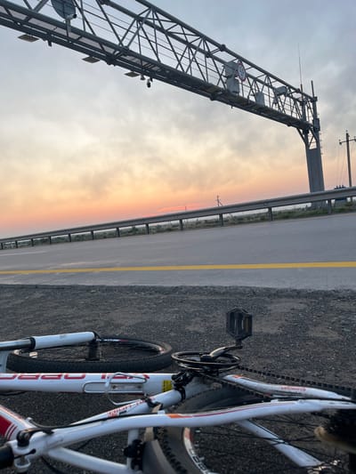 The image depicts a fallen white bicycle lying on a gravel shoulder beside a highway in Қоянды ауылдық округі, Kazakhstan. The bicycle is positioned horizontally, with its wheels facing upwards. The foreground is dominated by the bicycle's frame, pedals, chain, and partially visible wheels. In the background, the highway stretches out with a yellow dividing line. Overhead, a large metal gantry structure spans the highway, equipped with traffic lights, cameras, and signage, including a speed limit sign indicating 55. The sky above displays a gradient of sunset colors, ranging from pale orange near the horizon to a muted grey higher up, with wispy clouds. The environment appears arid, with sparse vegetation visible beyond the guardrail. No people are present in the scene.