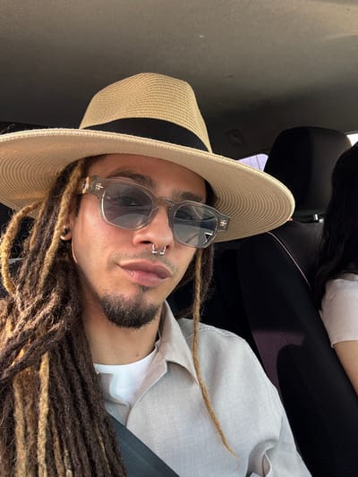A close-up portrait, oriented vertically, captures a person from the chest up inside what appears to be a vehicle. The subject has long, brown dreadlocks with lighter highlights, a short beard, and a mustache. They wear a light-colored, possibly off-white or light grey, collared shirt. A wide-brimmed straw hat with a black band covers part of their head, and they are wearing stylish, translucent grey sunglasses. The person has multiple facial piercings, including a silver septum ring and a ring on their left nostril. They look directly at the camera with a neutral expression.
Bright, natural sunlight illuminates the scene, casting highlights on the dreadlocks, face, and hat, suggesting a sunny day. In the background, the interior of the vehicle is visible, including a dark headrest and a light-colored side panel. Part of another person, wearing a white top and black clothing, is partially visible in the upper left background. The overall impression is a casual, personal shot taken while traveling, set in Corralejo, Spain. No visible text is present in the image.