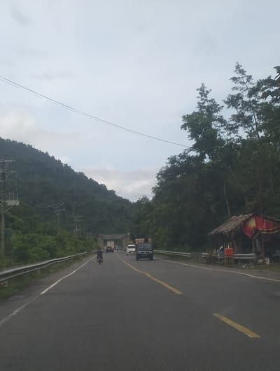 The image depicts a road scene in Seungkomulat, Indonesia. The road is paved and has yellow lane markings. There are vehicles on the road, including a motorcycle, cars, and trucks. On the right side of the road, there is a small roadside stall with a thatched roof and red and yellow banners. The stall appears to be selling goods. In the background, there are lush green hills covered in trees. Power lines and utility poles are visible on the left side of the road. The sky is overcast.