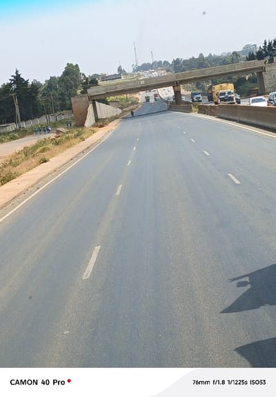 A highway stretches ahead, bisected by white dashed lines, leading towards an overpass. Beyond the overpass, a variety of vehicles including cars and trucks fill the road. Vegetation flanks the road on the left, while the right side features a concrete barrier. The image captures a sunny day, with some shadows cast on the road surface in the foreground. The sky is a bright blue.