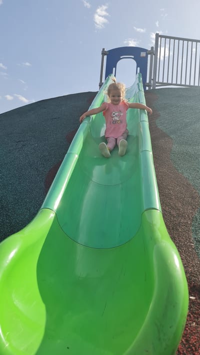 A young girl is riding a green slide in a playground in Kiryat Gat, Israel. She is wearing a pink shirt with a unicorn graphic and pink pants. The slide is located on a playground with a blue and grey structure. The ground is covered in a green and brown padded surface. The sky is blue with scattered clouds.