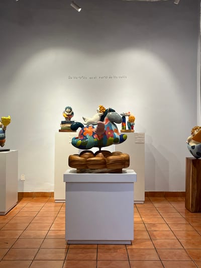 An interior shot displays artwork in a gallery. The art is mounted against a stark white wall, adorned with a faint inscription in Spanish. Sculptures and statuettes, vibrant with color and intriguing shapes, are displayed on pedestals. The floor is tiled with warm, terracotta squares, while spotlights cast shadows on the wall. The overall composition is clean and minimalist.