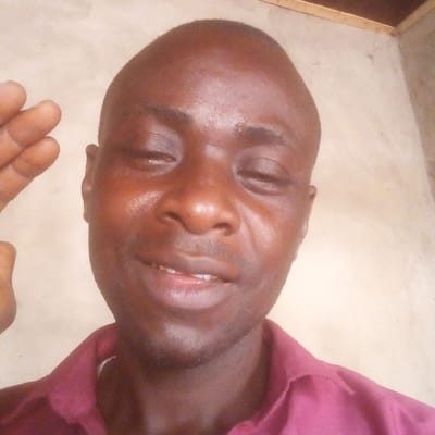 The image displays a close-up, chest-up portrait of an adult male, taken against a plain, light-colored background. The male subject has dark skin, a bald head, and appears to be smiling with his eyes partially closed. His face shows a noticeable sheen. He is wearing a maroon collared shirt. His right hand is partially visible on the left side of the frame, raised with fingers slightly separated, forming an open-hand gesture. The background wall is uniform in color, with a darker, textured area visible in the upper right quadrant, possibly indicating a structural corner or ceiling transition. This scene is located in the city of Ibreda, Nigeria.