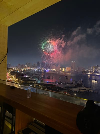 Night view from a high vantage point shows colorful fireworks exploding over a city skyline and a body of water with boats. The scene is captured from an elevated area with a wooden railing and a partially visible object in the foreground. City lights illuminate the cityscape below the fireworks, and reflections shimmer on the water. The sky is dark, enhancing the brilliance of the pyrotechnic display.