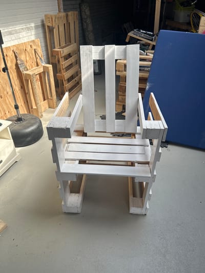 A white, unfinished chair made from pallet wood sits in the center of the frame. The chair has a high back with three vertical slats and armrests. The seat is made of horizontal slats. The chair is constructed from rough, unvarnished wooden pallets, some of which are still visible in their original state. The chair's construction suggests it is a DIY project. To the left of the chair, a black, weighted base of what appears to be a floor lamp or a stand is visible, along with a light-colored cabinet. Behind the chair, several more wooden pallets are stacked, some upright and some on their sides. A large, bright blue tarp or mattress covers a significant portion of the background to the right. The floor is a light gray concrete. The scene appears to be in a workshop, garage, or outdoor covered area, possibly a carport, given the presence of unfinished materials and tools that might be implied by the context of pallet furniture construction. The location is in Lisse, Netherlands.