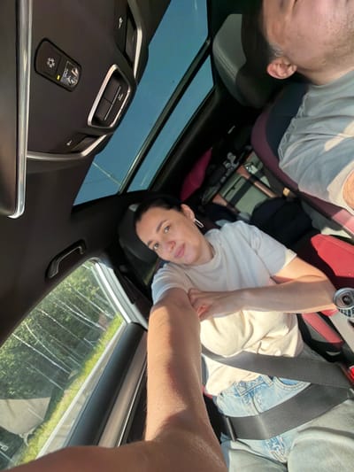 A first-person perspective shot captures two individuals inside a car, the sun roof is open, revealing a clear blue sky and a glimpse of trees and foliage outside. The driver, a woman with dark hair, is wearing a light-colored t-shirt and jeans and is looking towards the camera with a slight smile. She has a silver earring in her left ear. A seatbelt crosses her chest. In the passenger seat, a man with short hair is partially visible, wearing a light-colored t-shirt. The interior of the car is dark, with visible buttons and controls on the ceiling. The car's seats are upholstered in red and black. The overall lighting suggests it is daytime. The image is assumed to be taken in Uvarovo, Russia, based on the prompt.