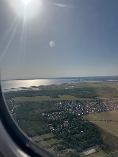This is an aerial view from an airplane window looking out over a suburban area and a body of water. The sun is prominently visible in the upper left corner of the image, casting a bright glare and lens flares across the sky. Below the sky, a large body of water stretches across the horizon, its surface reflecting the sunlight. The water appears to be a strait or bay, with land visible on the opposite side. In the foreground, a town or village is spread out, characterized by numerous houses with red or dark roofs. Green vegetation, including trees and fields, surrounds the residential areas and separates them from the water. A road is visible cutting through the landscape. The image captures a bright, sunny day with clear skies, suggesting it was taken during daylight hours. Based on the provided context of Tårnby, Denmark, this view likely encompasses parts of the municipality, possibly including areas bordering the sea or Oresund, with the town of Tårnby or its surrounding residential developments visible from above.