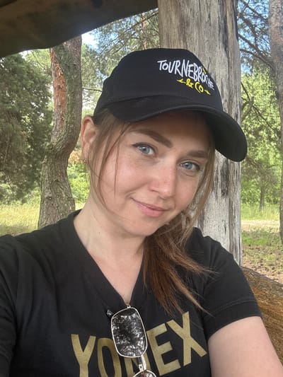 A selfie taken outdoors shows a woman wearing a black baseball cap with white and yellow text that reads "TOURNEBROCHE & CO". She has blue eyes and light brown hair. She is also wearing a black t-shirt with large gold lettering that reads "YONEX". Sunglasses hang from the neckline of her t-shirt. The background consists of trees and foliage, suggesting a natural or park-like setting. The lighting indicates it is daytime. The scene is likely located in Chmyrivka, Ukraine, given the context.