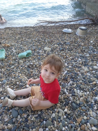 A young child, wearing a red t-shirt and tan shorts, sits on a pebble beach. The child has fair hair and is looking towards the camera with a curious expression. Their hands are reaching out, holding small twigs. The child is wearing light-colored sandals.
To the left of the child, the shallow, wavy water of the sea laps against the shore. The water is clear and reflects the light, creating a bright, serene atmosphere. A few pieces of driftwood and some large white and grey stones are visible along the water's edge, partially submerged. The rest of the scene is covered in a dense layer of small, smooth pebbles in various shades of grey, white, and brown, typical of a beach in Gwandra, Abkhazia.
Two light green Croc-style shoes are placed on the pebbles, one near the child's feet and another further down the beach. The overall impression is a peaceful day at the beach, with the child engaged in playing with natural elements. The lighting suggests it is daytime, likely with clear weather, given the brightness of the water and the scene. There is no visible text in the image.