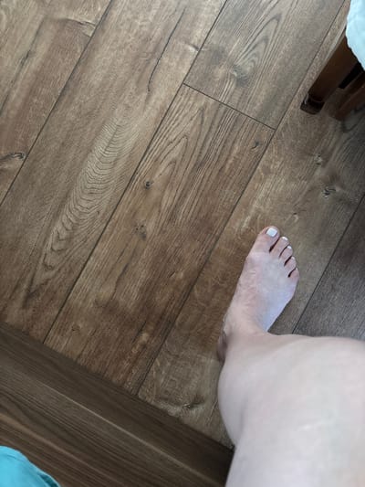 A close-up, top-down shot shows a person's bare foot with white-painted toenails resting on a wooden floor. The foot is positioned in the upper right quadrant of the image, with the toes pointing towards the left. The skin on the foot appears pale and shows some texture. The wooden floor is composed of planks with a brown, wood-grain pattern. The planks are laid diagonally across the image, creating a sense of perspective. In the upper left corner, a portion of what appears to be furniture with a dark wood finish is visible. The lighting suggests an indoor setting, likely during the daytime. There is no visible text in the image.