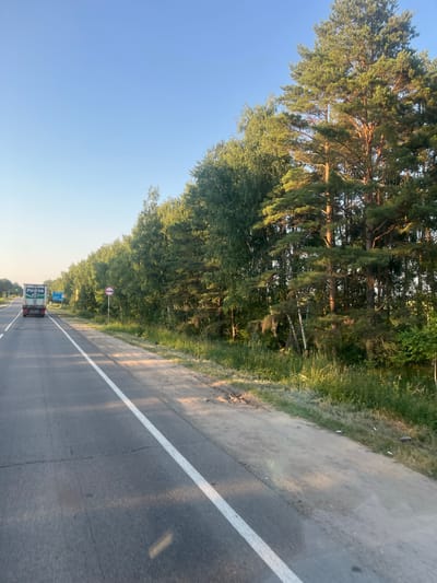 The image depicts a view from a vehicle traveling on a paved road through a wooded area in the settlement of Sovkhoza "Astapovo", Russia. The road has a white line marking the lane. On the left side of the road, a dense forest of pine and deciduous trees lines the verge. The sky is a clear, bright blue, indicating a sunny day. The time of day appears to be late afternoon or early evening, as suggested by the long shadows cast by the trees across the road.

In the distance, a white truck with a green logo and text is visible on the road. The logo on the truck appears to say "PULVA" in blue lettering. There are also blue road signs visible in the distance. A circular road sign with a red border and a white background, depicting a black symbol that appears to be a truck, is also present on the side of the road. The overall scene conveys a sense of travel through a rural, natural environment.