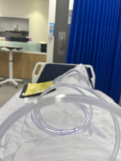 In a blurry, tilted shot, a clear oxygen tube dominates the foreground on a white patterned hospital bed sheet. A dark bed tray is partially visible on the bed. Behind the bed, a blue curtain hangs beside a gray wall with a silver handle and a blue label. Further back, a hospital room is visible with light-colored furniture, a door, and a framed picture. The scene evokes a sterile, clinical setting.