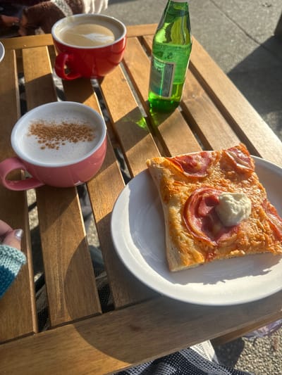 A square slice of pizza topped with prosciutto and a dollop of white cheese sits on a white plate. A green bottle lies horizontally, and two mugs filled with lattes are positioned beside it. One latte has latte art, while the other is sprinkled with cinnamon. All items rest on a wooden slatted table bathed in sunlight.