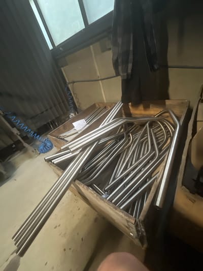 In an industrial setting, possibly a workshop or storage area, a wooden crate is filled with numerous metal pipes, many of which are bent into curved shapes. Some of these pipes are lying in a pile, with others extending out of the crate. Adjacent to the crate, a blue coiled hose is visible, connected to a larger coiled hose that runs along a ribbed, dark-colored wall. Above, a section of translucent roofing or ceiling panels allows diffused light to enter the space. A dark plaid garment hangs on the wall in the background. The floor appears to be concrete. The scene is located in Bogucin, Poland.