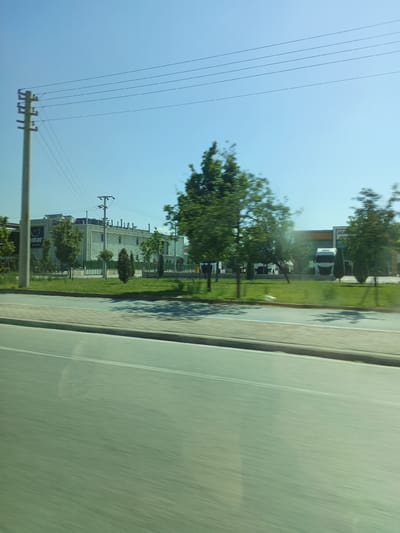 This media file captures a roadside view under a clear blue sky, suggesting a daytime setting with good weather. The scene is a commercial or industrial area in Karatay, Turkey.
Prominent in the foreground is a road with its lanes and a sidewalk, implying that the media was taken from a moving vehicle. Beyond the sidewalk, a strip of green grass is visible, dotted with trees and smaller bushes. In the mid-ground, several modern buildings are present. One building, partially visible on the left, has the text "binar" and "SÜT ÜRÜNLERİ" on its facade, indicating a dairy products company. Adjacent to it is another large building with a distinctive facade of horizontal slats and various pipes or vents on its roof. Further to the right, another building complex is seen, featuring large trucks parked in front of it. One truck, a white semi-trailer, is particularly noticeable.
A utility pole with multiple wires stretches across the upper portion of the frame, a common sight in many developed areas. The overall atmosphere is neutral and functional, typical of an industrial or commercial district. The clear sky and the visible greenery suggest a pleasant time of day, likely late morning or afternoon. The motion blur in the foreground indicates that the photo was taken while in transit, adding a sense of movement to the scene.