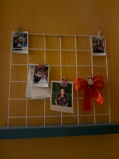 The image shows a white wire grid hanging on a textured yellow wall. Several photos are clipped to the grid. In the top left, a photo of three people, two adults and a child, is clipped with a green decorative clip. To the right, a photo of a person wearing a hooded jacket is clipped with a pink clothespin. In the middle left, a photo of five young adults is clipped to a piece of paper with a pink binder clip. The paper has a handwritten note that reads "to do". Below this, a photo of a young woman with dark hair, wearing a floral top, is clipped to the grid. To the right of this photo is a bright orange satin bow with a white circular embellishment in the center, clipped to the grid. The background is a textured yellow wall. A blue shelf is visible at the bottom of the image. The lighting suggests an indoor setting, possibly a home or a student dorm, with warm ambient light. The overall scene is a decorative display of personal photographs and a ribbon. No specific location in Kyiv, Ukraine, can be determined from the image.