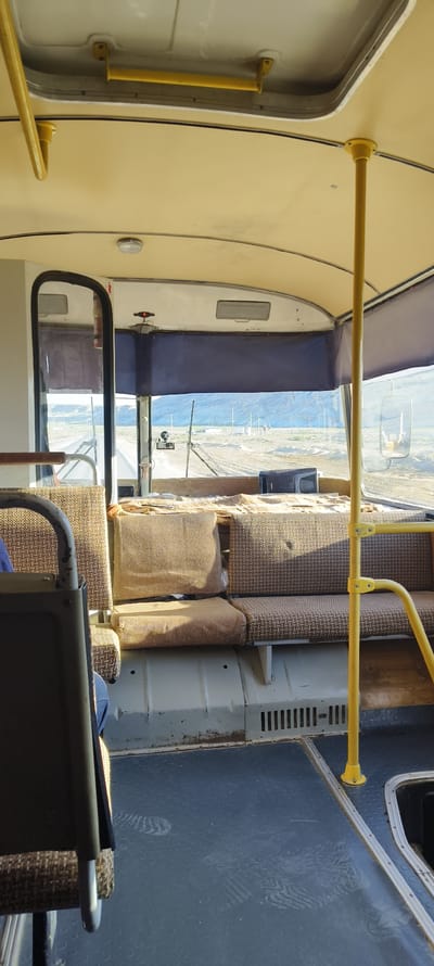 The image depicts the interior of an old bus. The seats are upholstered in a patterned, tan fabric, some of which appears worn and slightly torn. A yellow metal pole, common in buses for passengers to hold onto, is visible on the right side of the frame. The floor of the bus is a dark, textured material with some scuff marks and what appear to be footprints. In the background, through the bus windows, a sparse, dry landscape is visible, suggesting a rural or semi-arid environment. The lighting suggests it is daytime, with sunlight entering the bus. The overall condition of the bus interior suggests it is aged and possibly no longer in regular passenger service. This scene is consistent with the typical appearance of older public transportation or privately repurposed vehicles found in or around cities like Zhanaozen, Kazakhstan, where such older vehicles are still in use or have been maintained in varied conditions.