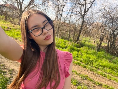 A young woman in a pink top and black eyeglasses poses outdoors, likely in a park or natural area. The ground is visible with grass and some bare patches. A canopy of bare trees is overhead. The scene appears to be in Blaskovo, Bulgaria, based on the location provided, but there are no specific landmarks that prove the location. The woman is looking down at the camera, her eyes are closed and her face appears relaxed. The background features the natural greenery of the ground and bare branches of the trees against a daytime sky.