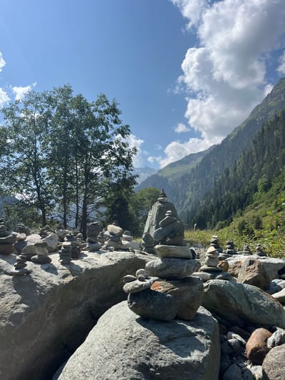 This image depicts a mountainous, rocky landscape in Reitlehen, Austria, under a clear sky with scattered white clouds. The foreground is dominated by numerous man-made rock cairns of varying sizes, composed of grey and earth-toned stones, balanced on larger boulders and loose rocky ground, likely a dried riverbed or glacial outwash. Sunlight casts distinct shadows across the scene, indicating a bright day. In the midground, several deciduous trees with green foliage are visible on the left. The background features steep, densely forested mountain slopes covered in dark green coniferous trees, rising towards the sky on the right. Distant mountain peaks are visible further back, appearing partially obscured by atmospheric haze. No individuals are present in the frame.