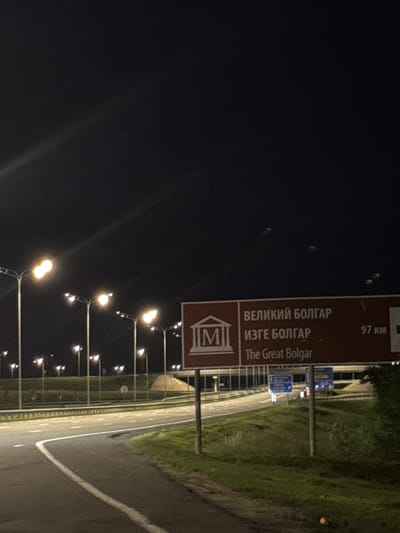 A night-time outdoor shot shows a road with streetlights illuminating the scene. On the right side of the road, a brown sign indicates "The Great Bolgar" with the distance "97 km" to the destination. The sign also features a building icon and text in Russian. Further in the background, more streetlights and traffic signs are visible. The road surface is asphalt, with white markings. The surrounding environment is grassy with some trees. The sky is dark.