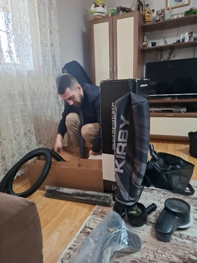 The image depicts an indoor residential setting in Zagore, Bulgaria. An adult male, appearing to be in his 20s or 30s with short dark hair and a beard, is crouched on a light-colored wooden floor. He is wearing a dark long-sleeved top and light brown trousers. His hands are inside an opened, light-brown cardboard box with internal dividers, suggesting an act of unpacking or assembly.
To the left of the man, a black corrugated hose, likely part of a vacuum cleaner, partially rests in and out of the box. To the man's right, a tall, black box stands upright, displaying "HOME CLEANING SYSTEM" and "KIRBY" in white text. Various black plastic vacuum cleaner attachments and components, some in clear plastic packaging, are scattered on a patterned rug in the lower right foreground. A black fabric bag, possibly for attachments, is also visible on the floor near the Kirby box.
The background features a window with a sheer, patterned lace curtain on the left. A light-colored two-door cabinet with a green and white Shrek figurine on top is situated against the back wall. To its right, an open shelving unit holds various decorative items, including framed pictures and small sculptures. Below the shelves, a black flat-screen television is positioned on a media console. The floor is light wood laminate or parquet, partially covered by a beige, brown, and white patterned rug.