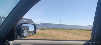 The image depicts a solar power plant in the city of Berezanka, Ukraine. The perspective is from inside a vehicle, looking out the side window and through the side mirror. Rows of blue solar panels are arranged in an open field under a clear blue sky. In the side mirror, a portion of the vehicle's interior and the surrounding landscape, including a building or structure, are reflected. The ground appears to be dry grass and dirt. A date and time stamp in the bottom right corner indicates "29.06.2026 11:20". There are no people visible in the image.