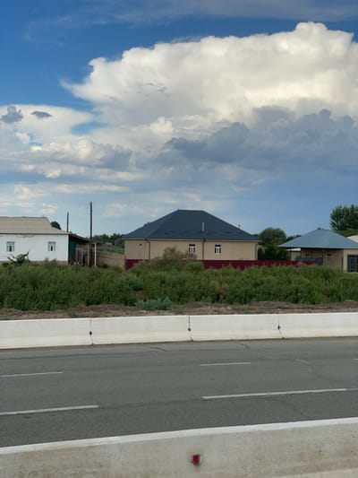 The image, taken in Qtay, Uzbekistan, depicts a roadside scene under a cloudy sky, suggesting it is daytime. The orientation of the image is portrait, with the bottom of the frame showing a concrete barrier bordering a two-lane asphalt road. The road has visible white lane markings. Beyond the road, a lush green area is filled with dense vegetation and tall grass.
Several buildings are nestled amongst the greenery. The most prominent is a large, modern-looking house with a dark grey, angled roof. It has a light-colored facade with several rectangular windows. To the left of this house, another building is partially visible, featuring a similar light-colored exterior and a dark roof. Further back, a simpler white building with a metal roof and windows is discernible, along with some older, less maintained structures that appear to be sheds or outbuildings, one with a green roof. A wooden utility pole stands near these structures.
The sky is dominated by dramatic, large cumulus clouds, interspersed with patches of blue sky. The light suggests it's a bright day, but the cloud cover creates some variation in illumination. There is no visible text within the image. The scene evokes a sense of rural or semi-rural environment, possibly on the outskirts of a town, with a mix of residential and possibly agricultural or utility structures.