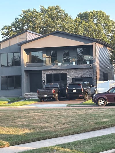 The image shows a modern, multi-story house with a dark gray exterior and large windows. In front of the house, there are two dark-colored SUVs and a dark pickup truck parked in the driveway. A portion of a burgundy sedan is visible on the right side of the frame. The house is surrounded by a well-maintained lawn. The scene appears to be in a suburban residential area. The image does not provide specific location details within Levittown, United States of America, other than it being a residential property. No people are visible in the image.
