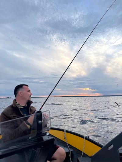 A man fishes from a boat on a body of water at sunset. The sky is filled with clouds, and the sun is setting on the horizon. The man is wearing a brown jacket and is holding a fishing rod. The boat is in the foreground, and the water extends to the horizon. The location is the city of Kostino, Russia.
