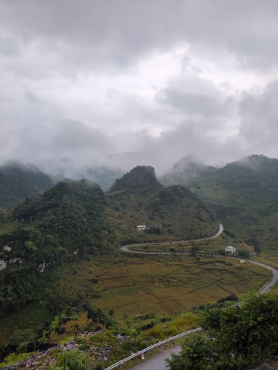 This image shows a scenic view of the mountainous landscape of Lũng Phìn Commune, Vietnam, under a cloudy sky. A winding asphalt road snakes through the hills, bordered by a metal guardrail and lush green vegetation. Terraced fields, likely planted with crops, are visible on the slopes, varying in color from green to a golden-brown hue, indicating different stages of growth or harvest. Several small, white houses are sparsely dotted along the hillsides and near the road. The mountains themselves are covered in dense green forests, with some peaks partially obscured by low-lying clouds and mist, creating a dramatic and atmospheric effect. The overall impression is one of rural tranquility and the majestic beauty of the Vietnamese highlands.