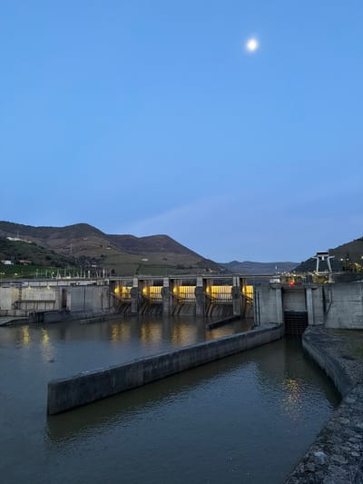The image depicts the Régua Dam (Barragem da Régua) on the Douro River, located near Lamego, Portugal, at twilight. A bright, circular moon is prominently visible in the clear, light blue sky above. The concrete dam structure extends across the mid-ground, featuring multiple spillway gates. The central five gates are illuminated by yellow-orange artificial lights from below, casting distinct vertical reflections onto the dark, calm river water. A concrete bridge runs horizontally across the top of the dam. To the right, a navigation lock system is visible, with a concrete channel leading into a lock chamber. A tall, white gantry crane structure stands on the far right bank above the lock, displaying red operational lights. Rolling hills covered with green vegetation form the background on both sides of the river. A dark, linear concrete barrier protrudes from the water in the foreground, parallel to the dam. No people are present, and no explicit actions or interactions are occurring.