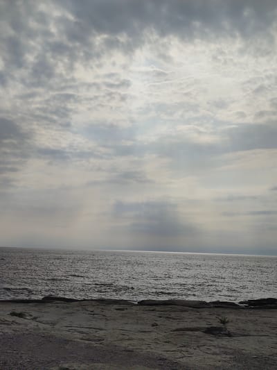 A wide, low-angle shot captures a cloudy sky over a body of water and a rocky shore. The sky is filled with a mix of dark gray and lighter, wispy clouds, with shafts of sunlight breaking through in some areas, creating a diffused, atmospheric light. The sea, stretching to the horizon, appears dark and slightly choppy, reflecting the somber mood of the sky. In the foreground, a rocky, uneven shore is visible, with scattered pebbles and some sparse, low-lying vegetation. The scene is static and devoid of any human presence or activity. The location is identified as Aqshuqyr, Kazakhstan.