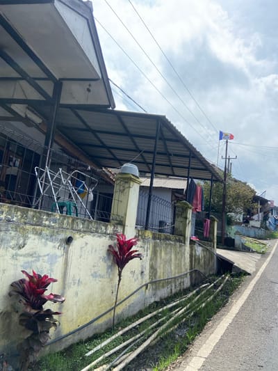 The image shows a residential street in the Kecamatan Cisarua area of Indonesia. The scene is dominated by a white concrete wall, which is covered in a layer of dirt and grime. There are a few red plants growing in the ground near the wall. The wall appears to be the front of a house, as there is a roof above it that appears to be an awning. The roof is supported by black metal beams. There is a fence in front of the wall, and a driveway that runs beside the wall and toward the left edge of the image. There are four large utility poles in the background, with a white flag hanging on one of them. The sky is a hazy blue. The street is paved in asphalt and extends toward the right edge of the image. There is a grass verge on either side of the street.