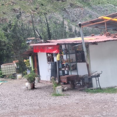 The image displays a small, single-story commercial building on a gravel lot in Ollantaytambo, Peru, set against a steep, vegetated mountainside. The structure features white walls and a red corrugated metal roof, with an additional roof extension on the right supported by metal poles and partially covered by a yellow tarp. A red banner bearing "Coca-Cola" logos is affixed to the front facade below the roofline.

To the left of the building, a large, white, grid-patterned intermediate bulk container (IBC tote) and several white plastic buckets are visible on the gravel ground. Potted plants are also present near the entrance. The front of the building includes an open entrance area where a metal shelving unit displays numerous bottles, likely beverages or provisions, and a yellow plastic bag hangs from it. A dark, vertical sign stands adjacent to the entrance.

To the right of the entrance, an outdoor cooking station is set up, comprising a metal stand with a griddle or grill top, containing wood fuel. Next to this, a table is covered with a striped or patterned cloth. A light-colored dog is partially visible standing near the building's entrance. The ground in the foreground and mid-ground consists of light-colored gravel, with small patches of green grass appearing on the far right.

The background is dominated by a rugged, verdant mountainside, densely covered with trees and exposed rock formations. A modern-style street lamp or light fixture is discernible high on the slope, and a distant, small building can be observed further up the mountain. No human individuals are visible in the scene, nor are any specific actions or interactions depicted.