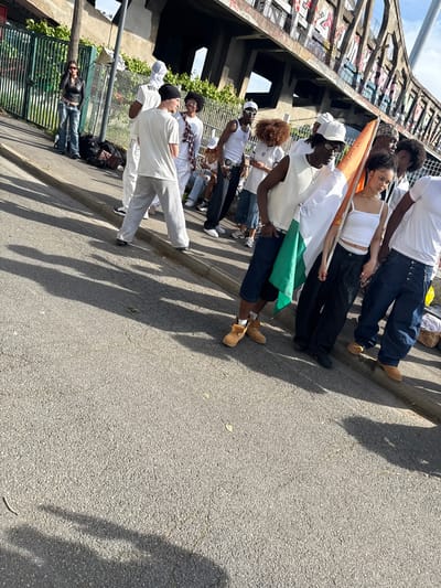 A group of approximately 10-15 young adults, predominantly of African descent, are captured walking or standing in a line on an asphalt surface in Charenton-le-Pont, France. The central figure holds a prominent flag of Côte d'Ivoire (Ivory Coast), characterized by its vertical orange, white, and green stripes, suggesting a cultural event, celebration, or procession.
Many individuals are dressed in casual attire, with a noticeable trend of wearing white tops such as t-shirts, tank tops, and hoodies, paired with jeans or trousers. One person is wearing a white hoodie with the text "BIG" visible on it. Some wear hats or caps, and one individual has on sunglasses.
To the left, a large, old concrete bridge or elevated structure, heavily covered in graffiti, forms a significant architectural backdrop. Green foliage and a metal fence line the path on the left side of the image. The scene is brightly lit by natural daylight, casting long, distinct shadows of the people onto the asphalt, indicating a sunny day. The sky above is clear with some scattered clouds. The overall mood appears relaxed and communal.
