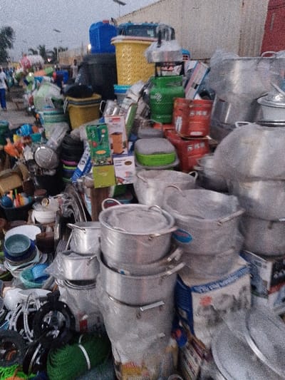 A cluttered outdoor market stall overflows with kitchenware. Stacks of aluminum pots dominate, interspersed with colorful plastic buckets, basins, and kitchen utensils. Gas cookers and various packaged goods are also visible. The scene appears busy, with blurred figures in the background, suggesting a bustling atmosphere. The light is soft, implying it's either early morning or late afternoon.
