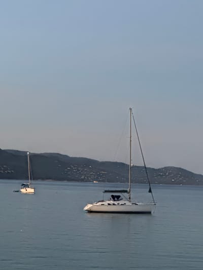 The image depicts a tranquil scene at sea, likely near Le Lavandou, France, during the twilight hours. Two sailboats are anchored in calm, blue water, with a hilly, verdant coastline dotted with lights in the background, suggesting a populated area. The sky is a pale, clear blue, transitioning to a lighter hue towards the horizon, indicating either early morning or late evening.

The sailboat in the foreground is larger and more detailed, featuring a white hull, a partially furled sail, and a dark canopy over the cockpit. Its mast is tall and slender, with rigging visible against the sky. The second sailboat, smaller and further in the distance, also has a white hull and a mast, appearing to be a racing or cruising sailboat. It is positioned to the left of the foreground vessel.

The water is smooth with subtle ripples, reflecting the dim light. The distant shoreline is characterized by rolling hills covered in vegetation, with numerous small, illuminated structures suggesting houses or buildings. The overall atmosphere is peaceful and serene. No people are visible in the image.