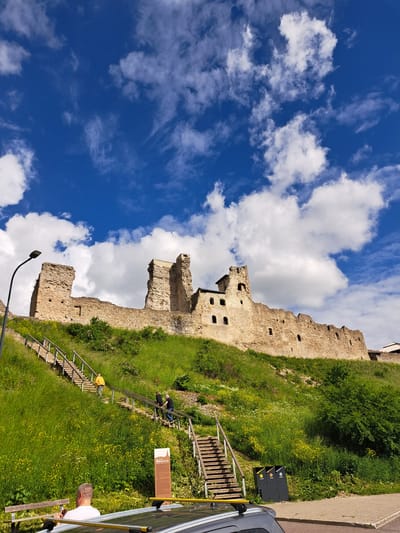 An eye-level, wide shot captures a sunlit view of the Rakvere Castle ruins in Rakvere, Estonia, against a bright blue sky filled with scattered white clouds. The ruins, constructed from stone, are perched atop a grassy hill, with portions of the walls and towers still standing. A long, wooden staircase with handrails winds up the side of the hill, leading towards the castle. Several people are visible on the stairs: one person in a yellow jacket is ascending, while two other individuals, dressed in dark clothing, are walking down. In the foreground, a portion of a dark-colored car is visible, with a man wearing a white t-shirt seated inside. To the right of the stairs, three dark trash bins are positioned near the base of the hill, adjacent to a paved area. The hillside is covered in lush green grass and dotted with small yellow wildflowers. A street lamp stands to the left of the staircase.