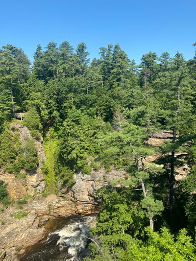 This vertical, high-angle shot captures a majestic waterfall cascading down a rocky cliff face, surrounded by a dense forest of evergreen trees. The water is a frothy white as it plunges into the dark river below. Sunlight dapples through the trees, illuminating parts of the lush green foliage and the rough, textured rock.
On the cliff face, a small wooden structure, possibly a viewing platform or a shelter, is nestled amidst the trees and rocks, adding a man-made element to the natural landscape. Several figures, appearing to be people, are visible standing on the edge of the cliff above the waterfall, seemingly enjoying the breathtaking view.
The sky is a clear, vibrant blue, suggesting a sunny day, likely in the late morning or early afternoon due to the bright lighting and clear visibility. The overall scene evokes a sense of awe and the raw beauty of nature, with the sounds of the roaring waterfall implied by the visual dynamism. No text is visible in the image. The location is the Town of Peru, USA.