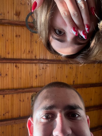 The image displays a low-angle shot of two individuals, with a wooden ceiling or wall serving as the background. The upper portion of the image features a female subject, positioned inverted, with her head partially obscured by her left hand. Her brown hair is visible, and her visible right eye (viewer's left) is brown and directed towards the camera. Her nails are long, pointed, and painted red with white and gold abstract designs. A silver-toned ring with small stones is visible on her ring finger. A dark, circular earring is partially visible through her hair on the left side of the frame.
The lower portion of the image features a male subject, right-side up, with the upper part of his head and face visible. He has short, dark brown buzzed hair. His brown eyes are open and directed towards the camera. Parts of his nose, cheeks, and left ear are visible.
The background consists of vertical wooden planks with two horizontal wooden beams. The wood has a warm, medium-brown finish, with visible grain and knots. The scene takes place in Staro Selo, Bulgaria.