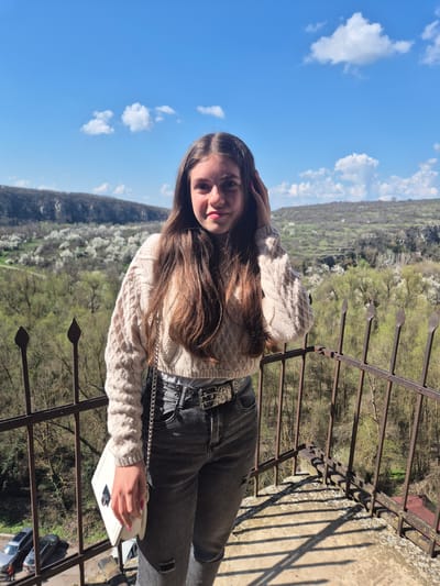 A young woman is standing on a balcony, facing the viewer. She has long brown hair and is wearing a beige knitted sweater, grey jeans with a belt, and is holding a white bag with a silver chain. Her right hand is touching her hair. She is situated against a backdrop of trees, a blue sky with clouds, and a metal railing. In the foreground, there are shadows and part of a car. The location is the Ivanovo, Bulgaria.